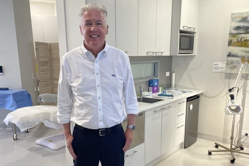 A smiling man in a white shirt and dark trousers stands in a modern clinic room with a treatment bed and kitchenette.